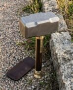 Damascus steel war hammer standing upright on its turned brass pommel on gravel beside a granite kerb and leather head cover