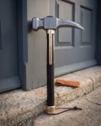 Damascus steel war hammer with crow beak pick and brass langets stood upright against a grey door, leather cover on the step