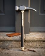 Damascus steel war hammer with dark hardwood haft and brass butt ferrule standing on a stone step, wrist thong at the base