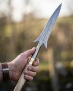 Damascus steel winged spear head with a brass ferrule on a pale ash haft, held upright in a hand with a blurred green field behind