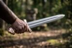 Damascus sword with brass lobed pommel and rune marked ricasso gripped in a hand, blade pointing along a shaded woodland path