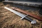 Damascus sword with five lobed pommel and black leather grip lying on dry straw beside a brown leather scabbard at a stone step