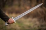 Damascus sword with five lobed pommel and runes on the guard held level in one hand, dry winter grassland behind