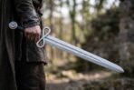 Damascus sword with figure of eight loop guard held point down at the side by a cloaked figure in a forest