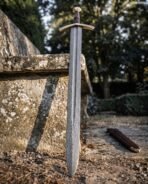 Damascus sword with random pattern blade and curved brass cross standing point down against a stone bench, scabbard behind