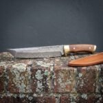 Damascus tanto blade with Elder Futhark runes etched along the spine, walnut scales and brass bolster resting on a lichen covered brick wall beside a tan leather sheath