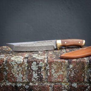Damascus tanto blade with Elder Futhark runes etched along the spine, walnut scales and brass bolster resting on a lichen covered brick wall beside a tan leather sheath