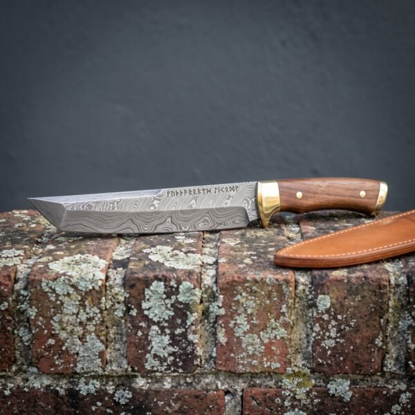 Damascus tanto blade with Elder Futhark runes etched along the spine, walnut scales and brass bolster resting on a lichen covered brick wall beside a tan leather sheath