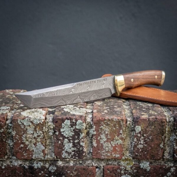Damascus tanto knife with etched blade and walnut handle laid flat on an old brick wall, tan leather sheath behind