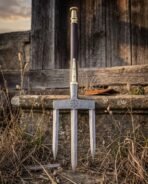 Damascus three prong trident standing upright in dry grass with dark haft and brass fittings against a timber wall