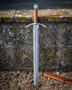 Damascus tree of life sword standing point down in gravel, folded steel blade with a central fuller, tan leather grip and a ricasso etched with Yggdrasil