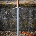 Damascus tree of life sword standing point down in gravel, folded steel blade with a central fuller, tan leather grip and a ricasso etched with Yggdrasil