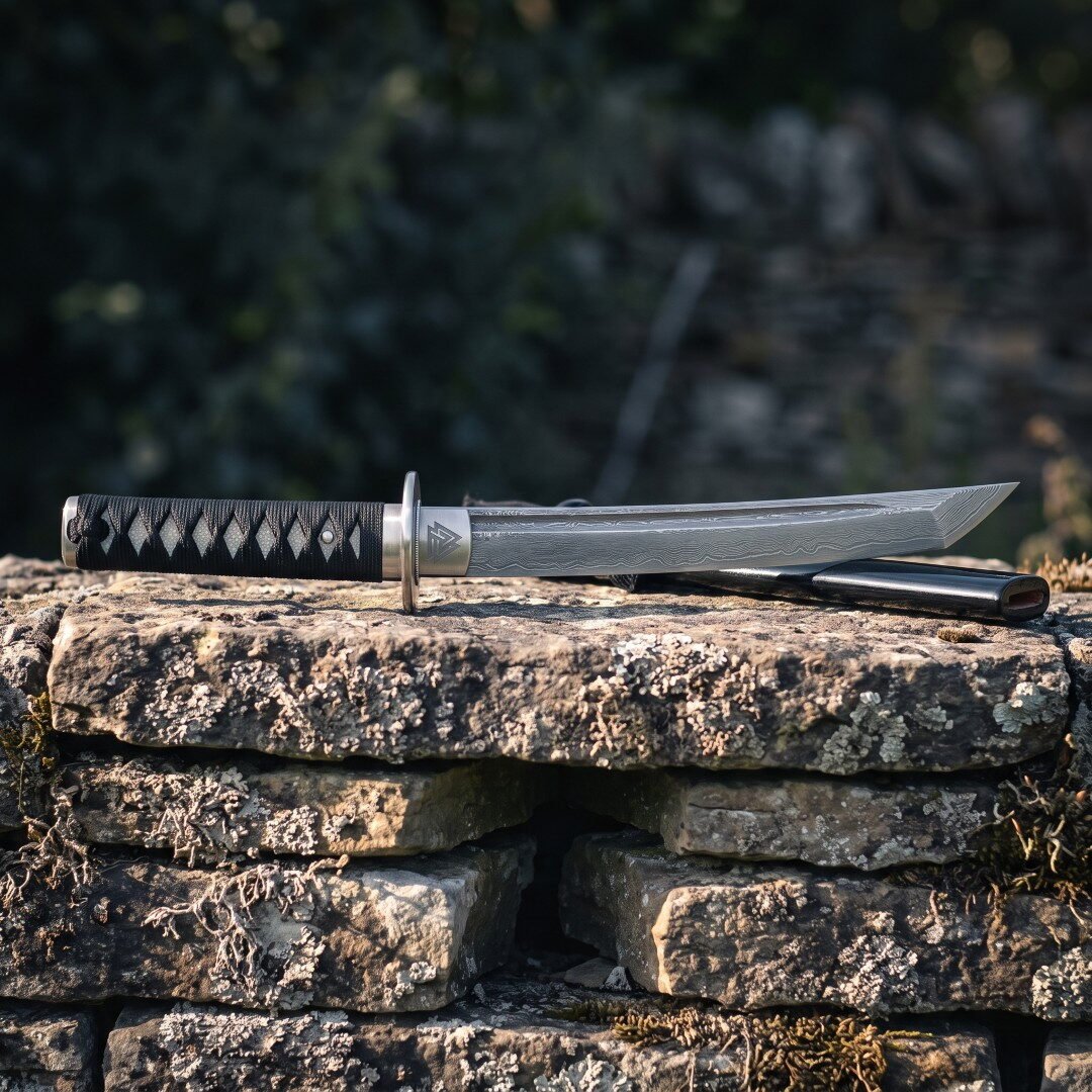 Damascus valknut tanto sword resting on a dry stone wall beside its black scabbard, showing the pattern welded blade, et Damascus valknut tanto sword resting on a dry stone wall beside its black scabbard, showing the pattern welded blade, etched Valknut, round steel tsuba and cord wrapped handle.