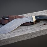 Damascus vegvisir kukri lying on weathered timber beside its brown leather sheath, showing the etched runic compass, deep recurved blade, brass bolster and black scales.