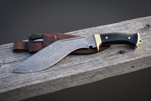 Damascus vegvisir kukri lying on weathered timber beside its brown leather sheath, showing the etched runic compass, deep recurved blade, brass bolster and black scales.