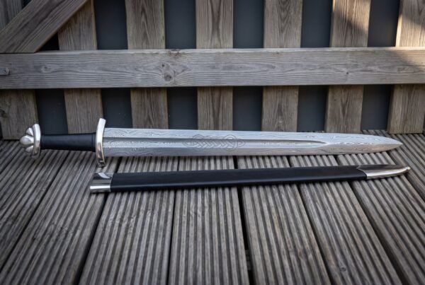 Damascus Viking broadsword lying on grey decking boards, showing a large etched knotwork panel on the blade, a black grip, brazil nut pommel and a black scabbard