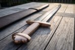 Damascus Viking sword hilt in close view showing the brass trilobate pommel and ribbed bone grip beside its brown leather scabbard on weathered decking