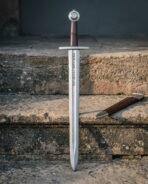 Damascus viking sword with Elder Futhark runes cut into the broad blade, leather wrapped grip and wheel pommel, standing on stone steps beside a timber scabbard