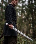 Damascus Viking sword with lobed steel pommel held point down by a man in a dark wool tunic in the forest
