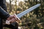 Damascus Viking sword with rune etched fuller and bronze toned guard held in a fist against green shrubs