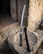 Damascus wakizashi standing beside its black lacquered scabbard on a sunlit stone trough, the curved blade and Celtic knot engraved collar set against a rough farm wall