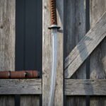 Damascus wakizashi sword with a ladder pattern blade, a brown leather wrapped handle and a knotwork tsuba, hanging against a weathered grey timber fence in bright sun