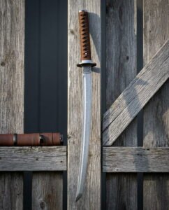 Damascus wakizashi sword with a ladder pattern blade, a brown leather wrapped handle and a knotwork tsuba, hanging against a weathered grey timber fence in bright sun