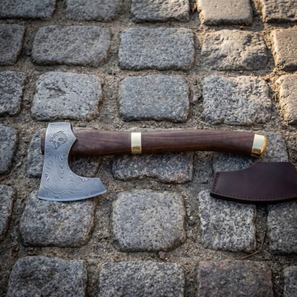 Damascus walnut haft bearded axe lying on cobblestones beside its brown leather edge sheath, showing the etched wolf head, swirling pattern welded head and brass collar.