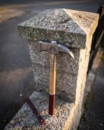 Damascus war hammer with etched ravens, ash haft and oxblood leather grip leaning on a granite gate post