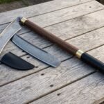 Damascus war scythe on a hardwood haft with brass collars laid across weathered decking beside a companion Damascus blade and its black leather sheath