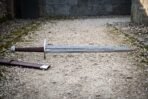 Damascus wheel pommel sword lying on pale gravel inside a stone gateway, showing a brown leather grip, straight steel crossguard and a cut knotwork panel near the ricasso