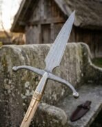 Damascus winged boar spear head with swept crossbar and brass knotwork ferrule on an ash haft by a stone bench