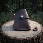 Dark brown leather axe sheath with a tooled Mjolnir panel and steel snap strap on a tree stump in the woods