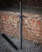 Dark finished black broadsword with a swept steel crossguard and wide diamond section blade standing point down on gravel by an old brick wall