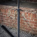 Dark finished black broadsword with a swept steel crossguard and wide diamond section blade standing point down on gravel by an old brick wall