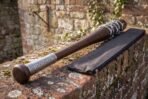 Dark hardwood baseball bat with barbed wire wrap and knotwork engraved collar resting on a mossy brick wall with its zip case