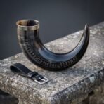 Dark ox horn drinking horn standing on its curve showing the full rune row and steel rim beside a black leather belt loop