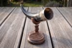 Dark ox horn drinking horn resting in a turned walnut cradle on a carved knotwork coaster, set on garden decking