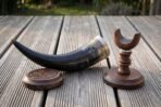 Dark ox horn drinking horn with stand in turned walnut, steel rim band and a carved knotwork coaster, photographed on weathered decking in low sun