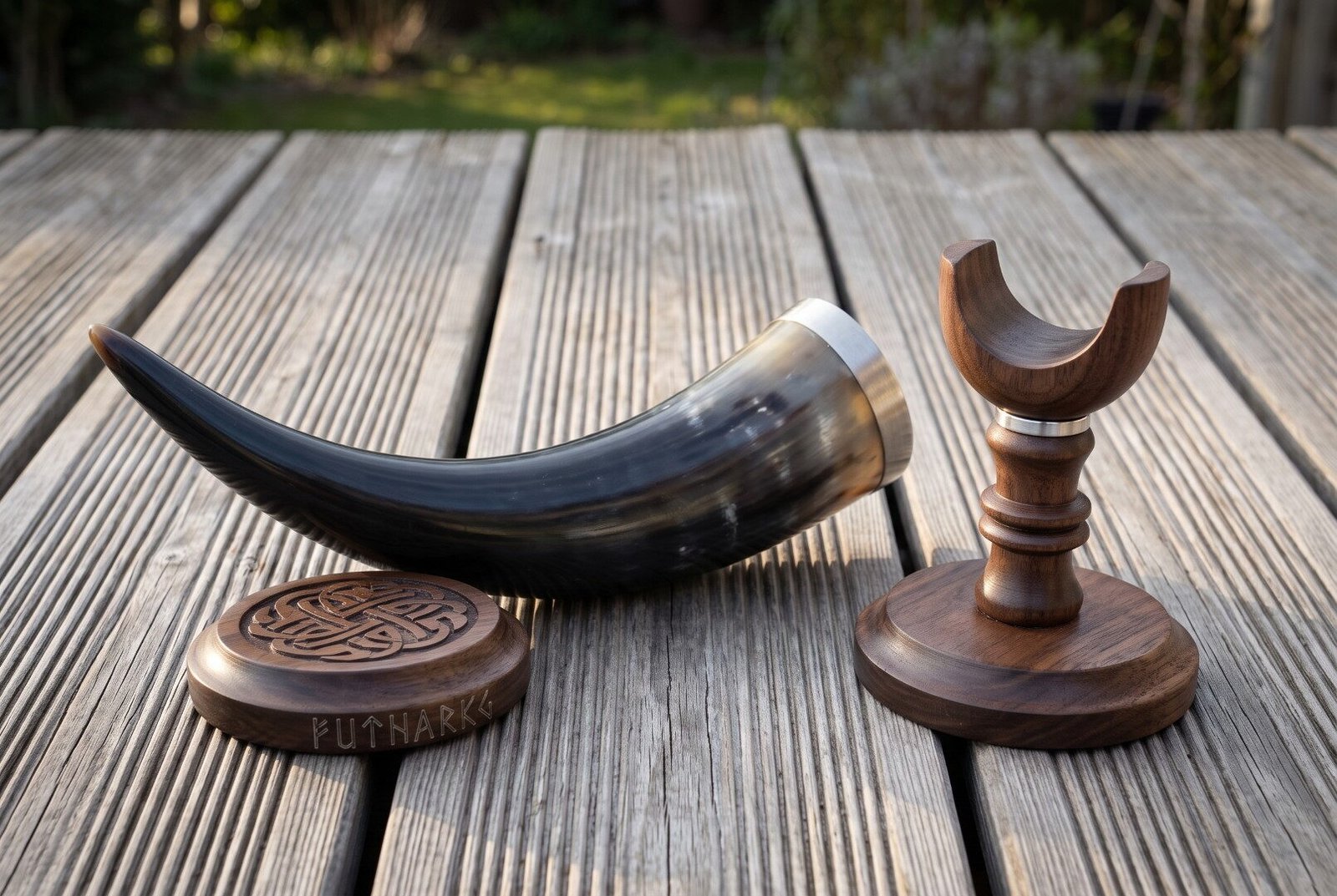Dark ox horn drinking horn with stand in turned walnut, steel rim band and a carved knotwork coaster, photographed on weathered decking in low sun