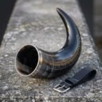 Dark ox horn drinking horn with steel rim and engraved runes along the curve, black leather belt loop with buckle on stone