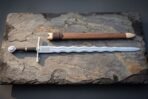 Disc pommel flamberge sword with a bright polished undulating wave blade, etched Celtic knot panel, black grip and brass mounted timber scabbard on a slate slab