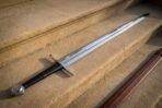 Disc pommel sword lying on sunlit sandstone steps with a fullered bright blade, black leather grip over cord risers and a brown leather scabbard alongside