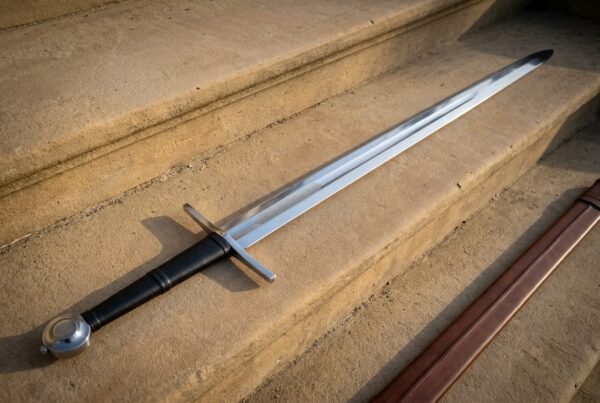 Disc pommel sword lying on sunlit sandstone steps with a fullered bright blade, black leather grip over cord risers and a brown leather scabbard alongside