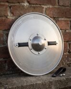 Front view of a dished polished steel round shield with etched knotwork border, riveted boss and two black leather grips against a brick wall