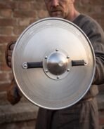 Dished steel round shield with etched knotwork border, riveted centre boss and black leather handles held by a man in a grey linen tunic