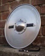 Dished steel round shield with etched knotwork border and riveted centre boss leaning against a brick wall, black leather strap on the ledge