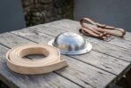 Domed steel shield boss beside a coiled natural leather rim binding and brown buckled straps on a weathered table, low side angle