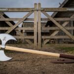 Double bladed Viking axe with twin polished 1095 steel crescent bits and an etched beast motif on a white ash haft standing before a timber longhouse gate