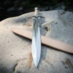 Double edged leaf shaped steel short sword with ribbed grip lying on a pale boulder across its natural leather sheath